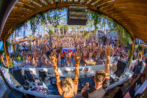 NFIX & CANDICE raising their hands behind a DJ booth under a wooden roof