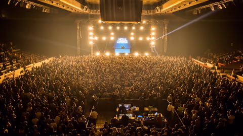 A packed indoor audience facing a brightly lit stage
